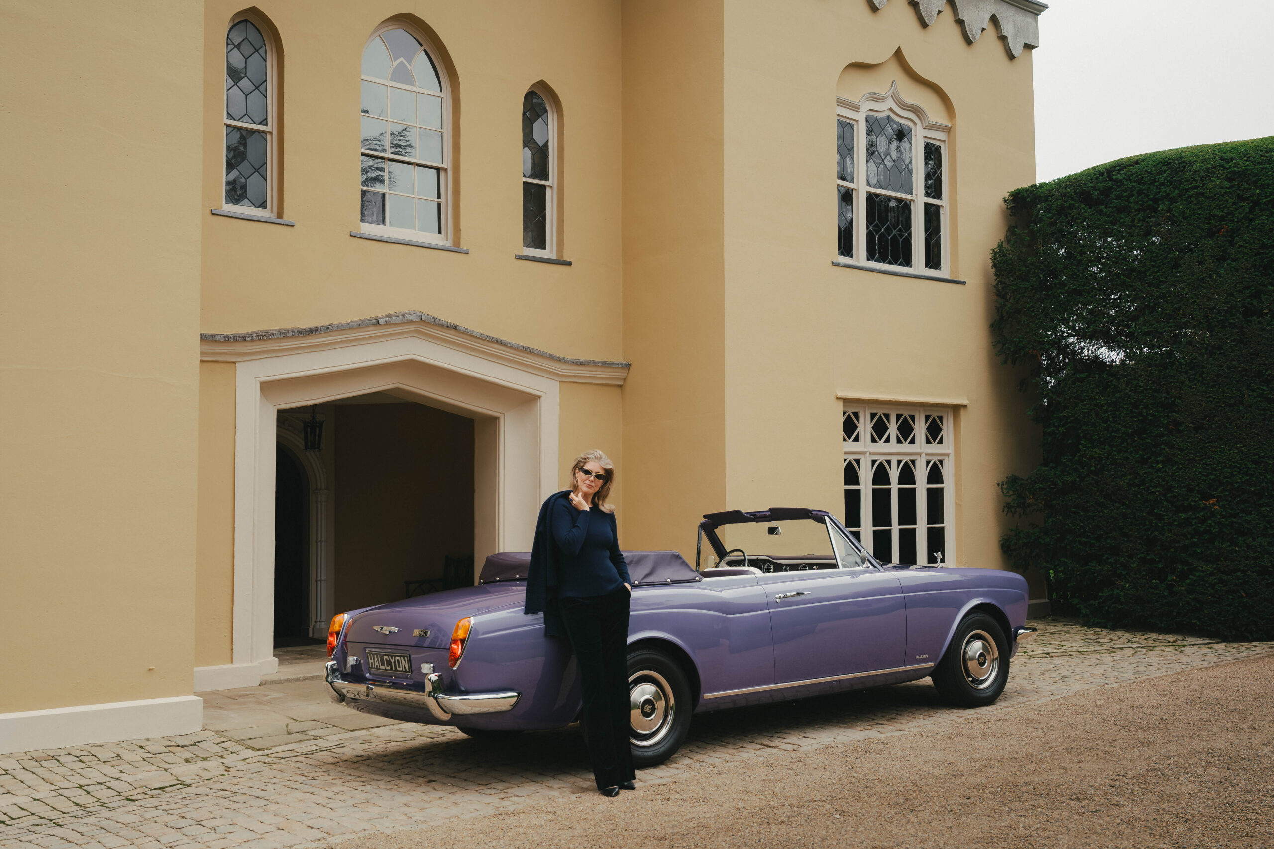 Woman in Renauld sunglasses and navy knitwear leaning against the rear of a lavender Halcyon remastered Rolls-Royce Corniche convertible outside the Gothick facade of Hampden House, Buckinghamshire, campaign production by 36 Studios