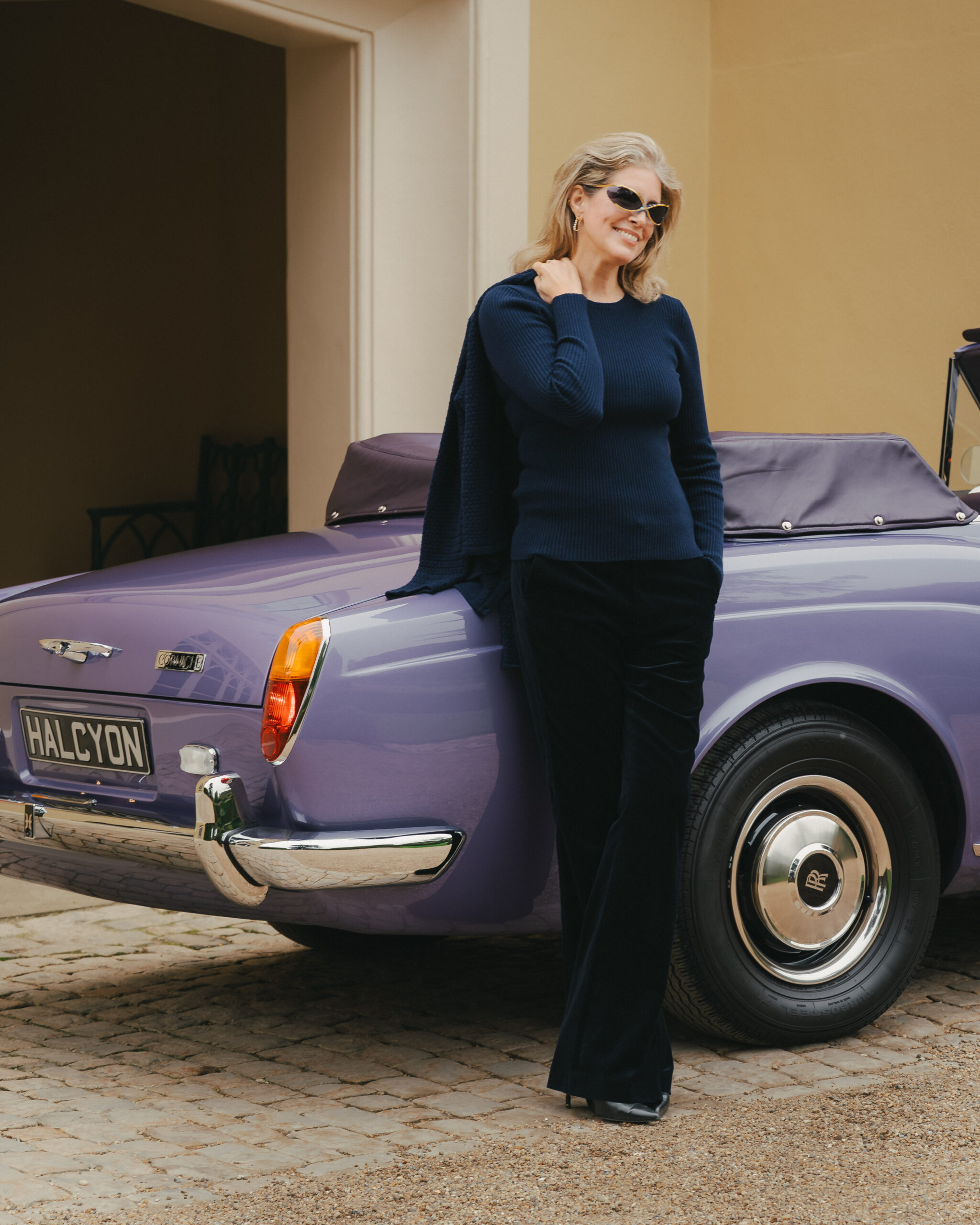 Smiling woman in gold-framed Renauld wraparound sunglasses, navy ribbed knit and black velvet trousers leaning against the rear wing of a lavender Halcyon remastered Rolls-Royce Corniche on cobbles at Hampden House, campaign production by 36 Studios