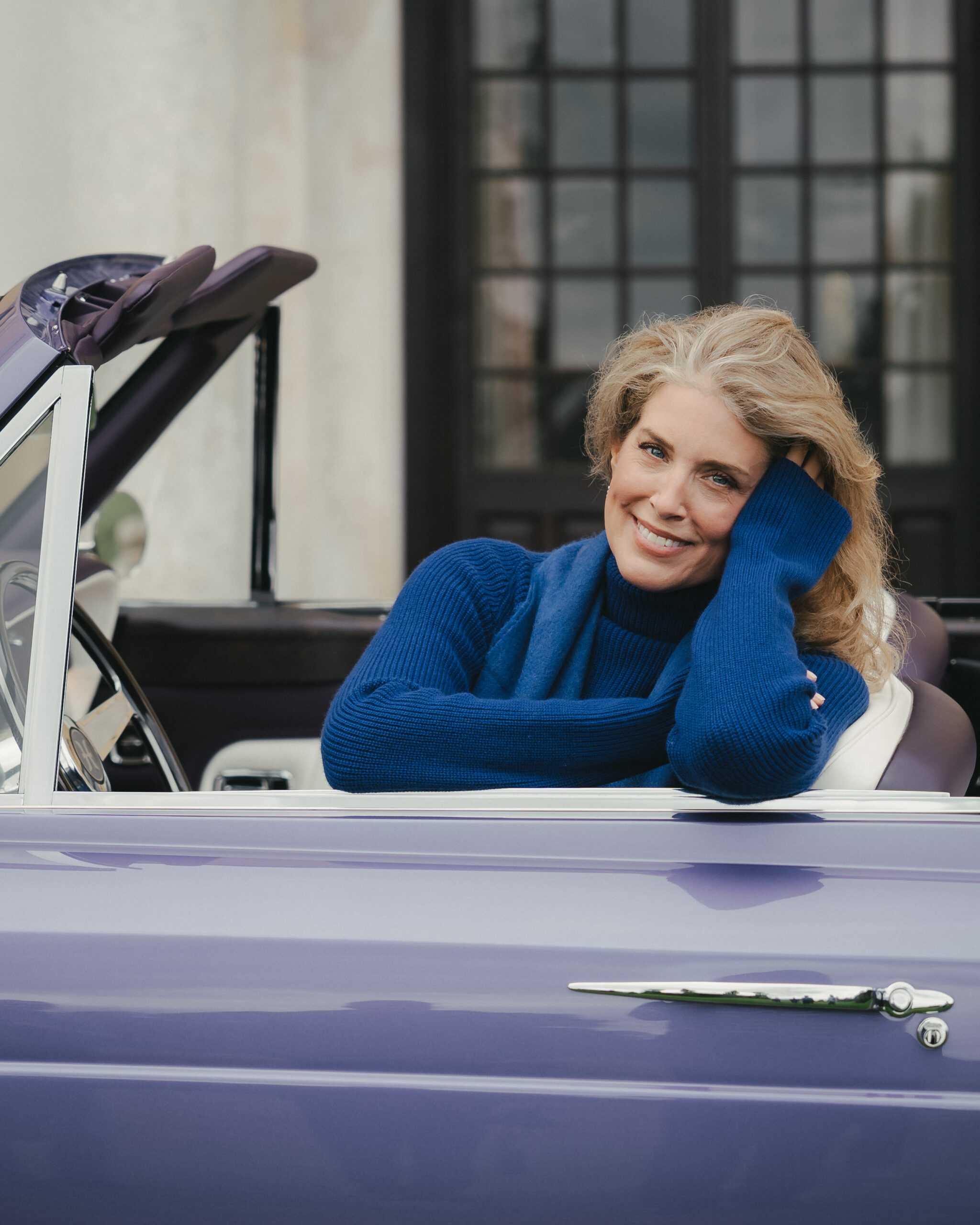 Smiling woman in a cobalt cashmere roll neck and scarf resting her head on her hand, seated in a lavender Halcyon remastered Rolls-Royce Corniche convertible with cream leather seats, Hampden House, campaign production by 36 Studios