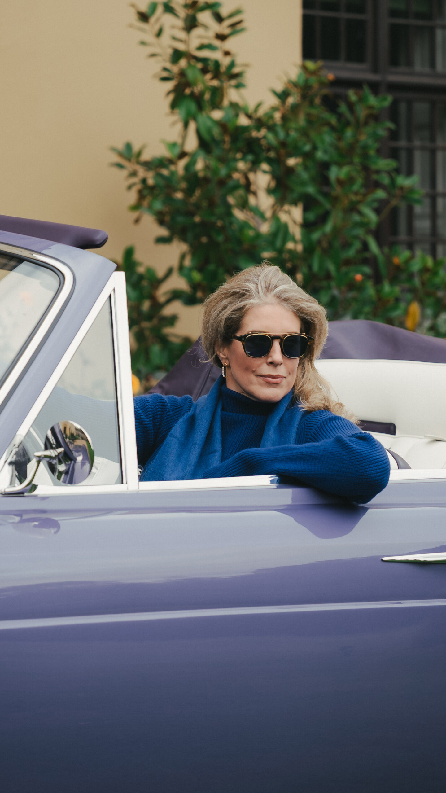 Woman in tortoiseshell Renauld sunglasses and a cobalt cashmere roll neck and scarf seated in a lavender Halcyon remastered Rolls-Royce Corniche convertible with cream leather interior, Hampden House, campaign production by 36 Studios