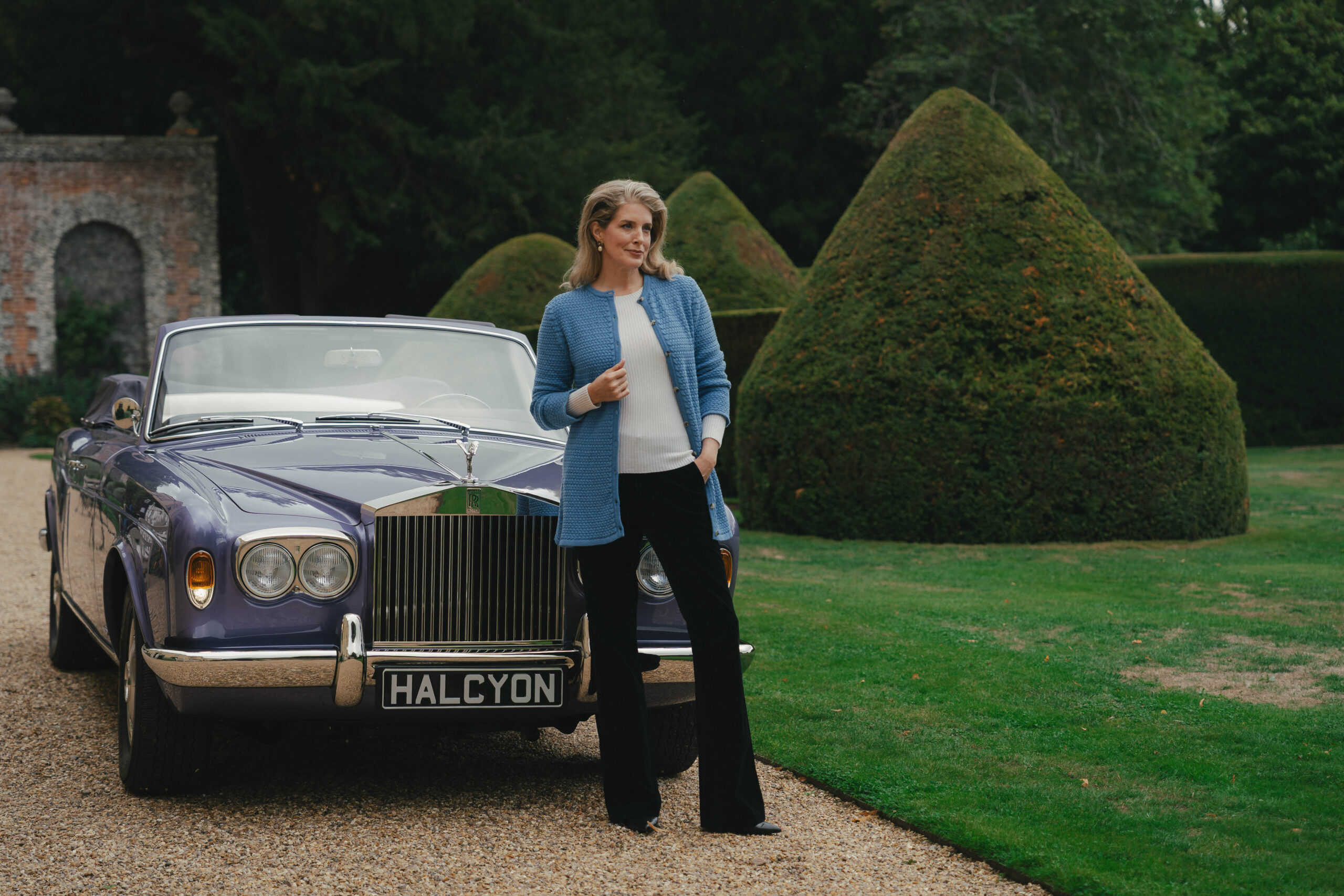 Woman in a blue cashmere cardigan leaning on the bonnet of a lavender Halcyon remastered Rolls-Royce Corniche convertible beside clipped yew topiary at Hampden House, Buckinghamshire, campaign production by 36 Studios