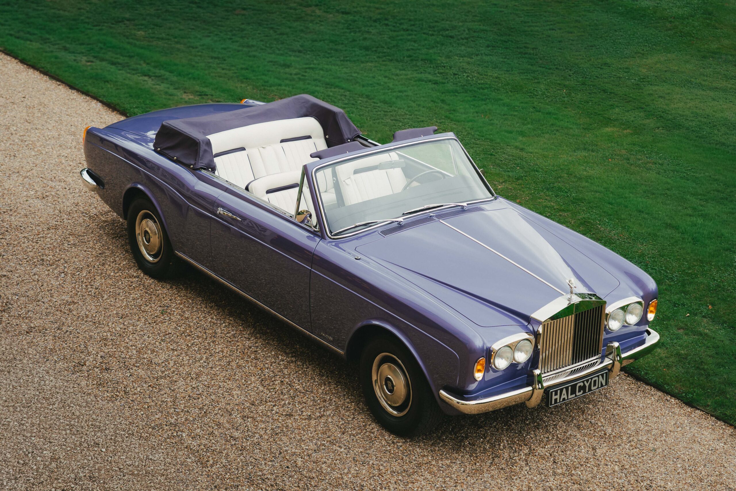 Elevated three-quarter view of a lavender Halcyon remastered Rolls-Royce Corniche convertible with hood folded and cream pleated leather seats, parked on a gravel drive beside a lawn at Hampden House, campaign production by 36 Studios