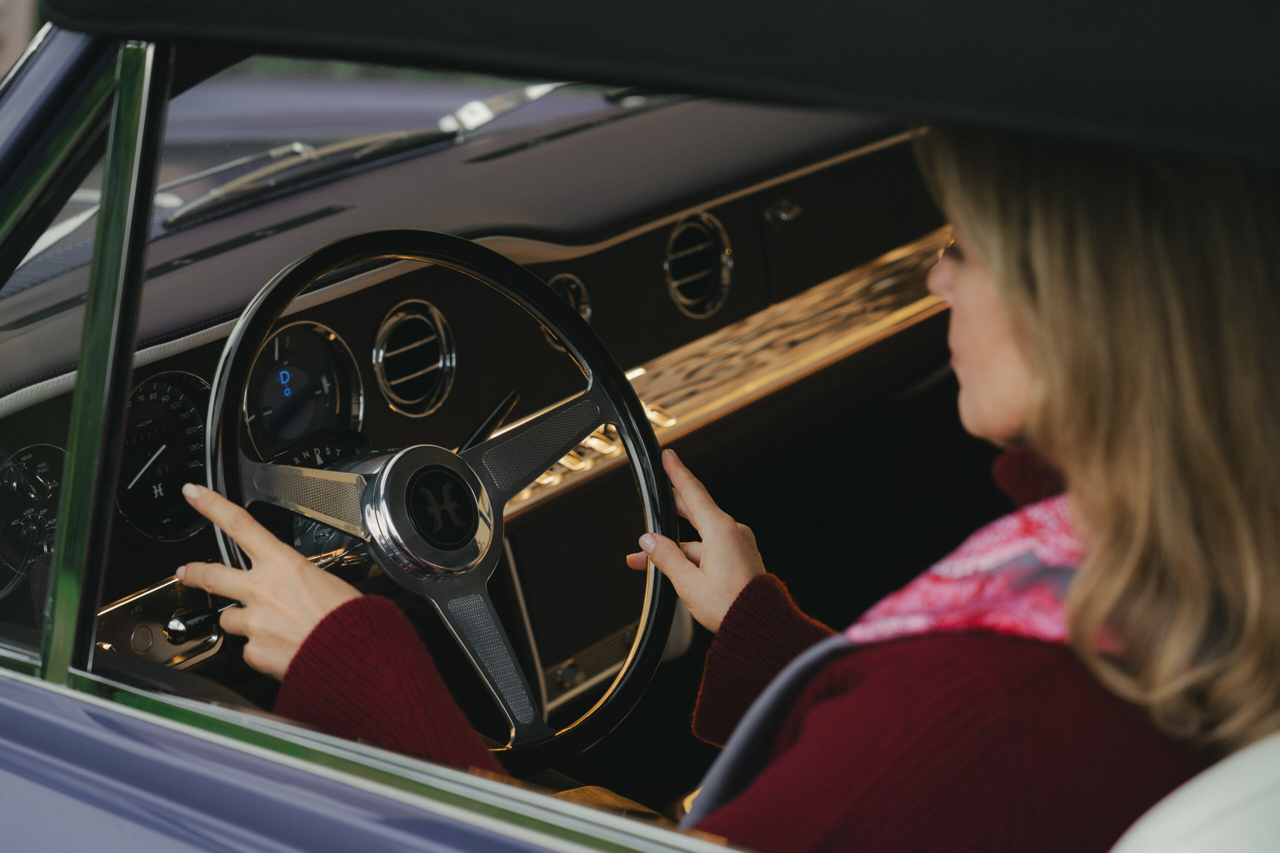 Over-the-shoulder view of a woman in a burgundy knit with hands on the polished steering wheel of a Halcyon remastered Rolls-Royce Corniche, Halcyon-branded hub, chrome vents and sculpted gold dashboard trim, campaign production by 36 Studios